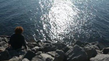 Arka manzara artı Afrikalı zenci kadın afro saçı gün batımında huzurlu bir deniz kenarında tatilin tadını çıkarıyor güneşli hava, kayalar, mavi okyanus suyu. Vücut pozitif çeşitlilik kavramı