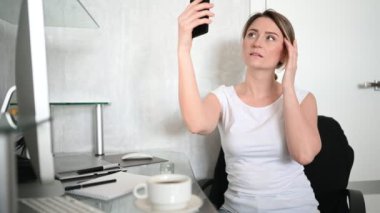 Teknoloji, video konferansı, sanal olay konsepti. Ev işyerindeki bilgisayarın önünde oturan mutlu genç kadın akıllı telefonla selfie ya da video görüşmesi yapıyor, telefonla konuşuyor..