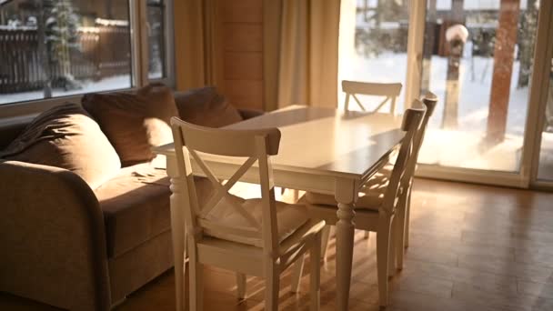 Ελαφρύς εσωτερικός σχεδιασμός του καθιστικού χώρα ξύλινο σπίτι στο σκανδιναβικό στυλ. Πανοραμικά παράθυρα, λευκό τραπέζι ομάδα τραπεζαρία και καρέκλες, άνετο μπεζ καναπέ. Χειμερινό τοπίο έξω από το παράθυρο. — Αρχείο Βίντεο