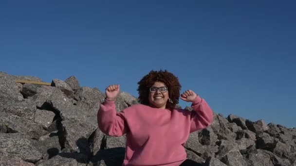 Schöne glücklich lächelnde plus Größe afrikanische schwarze Frau Afro-Haar in rosa Kapuzenpulli im Freien lachen tanzen genießen sonniges Wetter blauen Himmel. Körper Unvollkommenheit, Akzeptanz Körper positive Vielfalt Konzept — Stockvideo