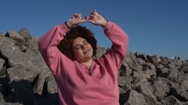 Schöne glücklich lächelnde plus Größe afrikanische schwarze Frau Afro-Haar in rosa Kapuzenpulli draußen genießen strahlendes sonniges Wetter und blauen Himmel. Körper Unvollkommenheit, Akzeptanz Körper positive Vielfalt Konzept — Stockvideo