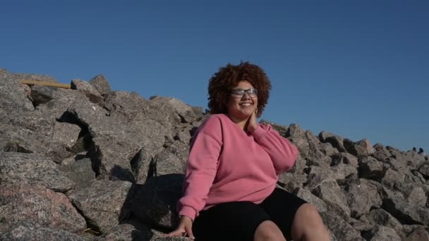 Mooie vrolijke glimlach plus size Afrikaanse zwarte vrouw afro haar in roze hoody buiten genieten van helder zonnig weer en blauwe hemel. Lichaamsimperfectie, acceptatie lichaam positief diversiteitsconcept — Stockvideo