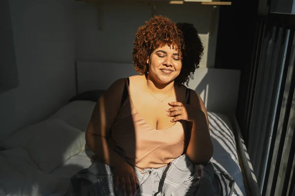 Indah melengkung ditambah ukuran Afrika hitam wanita afro rambut di tempat tidur dengan selimut kotak-kotak abu-abu nyaman desain interior. Sinar matahari. Ketidaksempurnaan tubuh, penerimaan tubuh, tubuh konsep positif dan keragaman — Stok Foto
