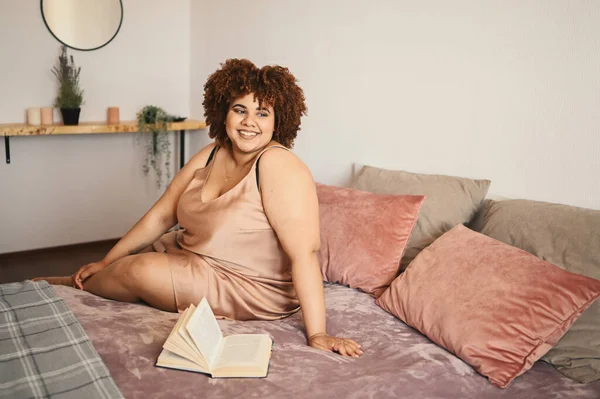 Linda curvilínea plus size Africano preto mulher afro cabelo deitado na cama em pó de seda rosa vestido aconchegante quarto design de interiores. Imperfeição corporal, aceitação corporal, corpo positivo e conceito de diversidade — Fotografia de Stock