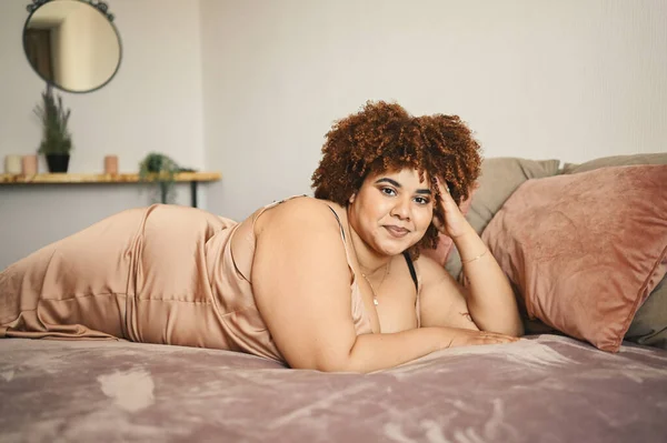 Indah melengkung ditambah ukuran Afrika hitam wanita afro rambut berbaring di tempat tidur dalam bubuk sutra merah muda gaun nyaman desain interior. Ketidaksempurnaan tubuh, penerimaan tubuh, tubuh konsep positif dan keragaman — Stok Foto