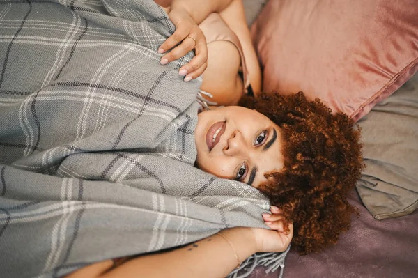 Indah melengkung ditambah ukuran Afrika hitam wanita afro rambut berbaring di tempat tidur dengan selimut kotak-kotak abu-abu nyaman desain interior. Ketidaksempurnaan tubuh, penerimaan tubuh, tubuh konsep positif dan keragaman — Stok Foto
