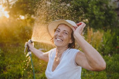 Mutlu yaşlı bir kadın yaz bahçesinde çiçekleri hortumla sularken eğleniyor. Arka planda bir damla su. Çiftçilik, bahçıvanlık, tarım, yaşlı insanlar. Çiftlikte organik sebzeler yetiştiriyor.