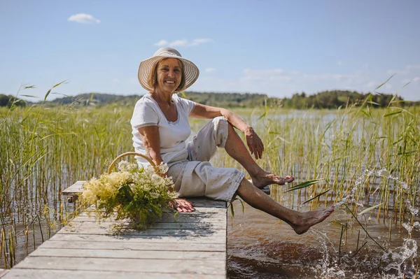 Feliz anciana sonriente con sombrero de paja sentada en el muelle de madera junto al lago con flores en la cesta. Agricultura, jardinería, agricultura, ancianos jubilados. País verano descanso —  Fotos de Stock