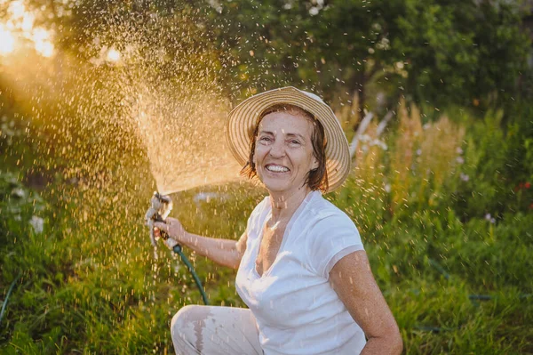 Wanita tua yang bahagia memiliki tanaman berair menyenangkan dengan selang di taman musim panas. Tetes air di backlight. Pertanian, berkebun, pertanian, orang-orang tua. Menanam sayuran organik di peternakan — Stok Foto