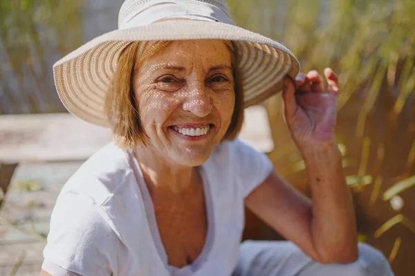 Foto close up dari wanita tua tersenyum bahagia dengan topi jerami duduk di dermaga kayu oleh danau. Pertanian, berkebun, pertanian, pensiunan orang tua. Istirahat musim panas Stok Lukisan  