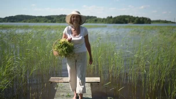 Glad leende äldre äldre kvinna i halmhatt promenader längs träpir vid sjön med blommor i korgen. Jordbruk, trädgårdsskötsel, jordbruk, pensionärer. Sommarvila i landet — Stockvideo