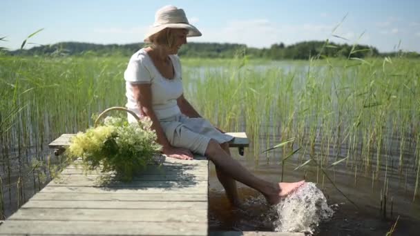 Щаслива усміхнена літня старша жінка в солом'яному капелюсі сидить на дерев'яному пірсі біля озера з квітами в кошику. Сільське господарство, садівництво, сільське господарство, люди похилого віку. Заміський літній відпочинок — стокове відео