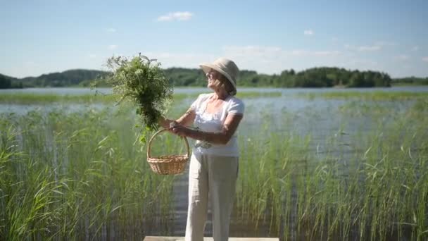 Щаслива усміхнена літня старша жінка в солом'яному капелюсі, що стоїть на дерев'яному пірсі біля озера з квітами в кошику. Сільське господарство, садівництво, сільське господарство, люди похилого віку. Заміський літній відпочинок — стокове відео
