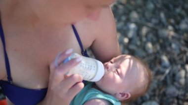 Yeni doğmuş, tatlı bir erkek çocuğu olan genç bir anne, onu kollarına alıyor, güneşli bir günde dışarıda süt şişesiyle sarılıyor ve besliyor. Sağlıklı çocuk, hastane kavramı ve mutlu annelik. Çocuk odası.