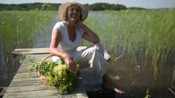 Glad leende äldre äldre kvinna i halmhatt sitter på träpir vid sjön med blommor i korgen. Jordbruk, trädgårdsskötsel, jordbruk, pensionärer. Sommarvila i landet — Stockvideo