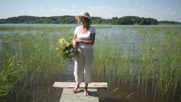 Glücklich lächelnde Seniorin mit Strohhut, die mit Blumen im Korb an der Seebrücke entlang spaziert. Landwirtschaft, Gartenbau, Landwirtschaft, Rentner. Sommerpause auf dem Land — Stockvideo