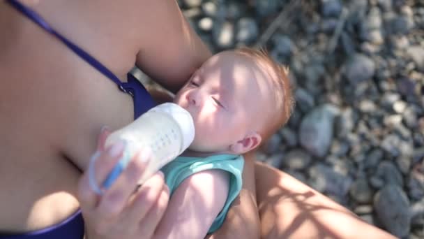 Yeni doğmuş, tatlı bir erkek çocuğu olan genç bir anne, onu kollarına alıyor, güneşli bir günde dışarıda süt şişesiyle sarılıyor ve besliyor. Sağlıklı çocuk, hastane kavramı ve mutlu annelik. Çocuk odası. — Stok video