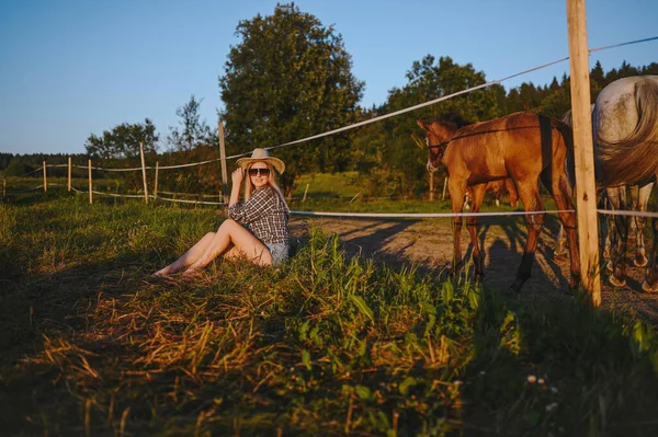 Hermosa mujer rubia joven vestido estilo safari en sombrero y camisa a cuadros posando con caballos pura sangre en la granja en el campo de atardecer al aire libre en el campo. Caballos detrás de una valla eléctrica energizada. —  Fotos de Stock