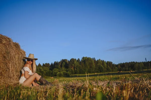 Młoda piękna blondynka rolnik kobieta w białym stroju, okulary przeciwsłoneczne kowbojski kapelusz i buty pozowanie o zachodzie słońca na polu z stogów siana na wsi. Koncepcja zbiorów. Modelka na zewnątrz. — Zdjęcie stockowe