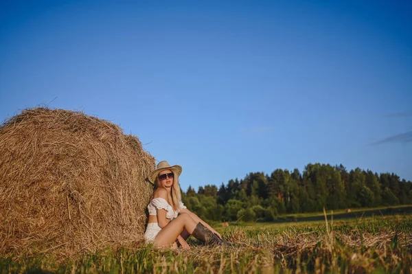 Joven mujer granjera rubia hermosa en traje blanco, gafas de sol sombrero vaquero y botas posando al atardecer en el campo con pajar en el campo. Concepto de cosecha. Modelo de moda al aire libre. —  Fotos de Stock