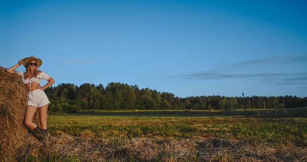 Joven mujer granjera rubia hermosa en traje blanco, gafas de sol sombrero vaquero y botas posando al atardecer en el campo con pajar en el campo. Concepto de cosecha. Modelo de moda al aire libre. —  Fotos de Stock