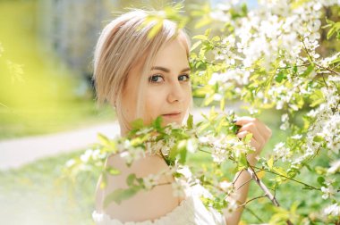 Genç, mutlu, gülümseyen sarışın kadın, çiçek açan bahar bahçesinde beyaz çiçeklerle poz veriyor. Çiçek açan elma ya da kiraz ağacı. Doğal kozmetik, gençlik, sağlıklı cilt ve dişler kavramı.