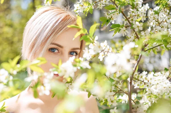 푸른 눈을 가진 예쁜 금발의 젊은 여성을 가까이 다가가서 하얀 꽃이 피는 봄 정원에 포즈를 취한다. 꽃이 피는 사과나 벚나무. 천연 화장품, 젊음, 건강 한 피부와 치아의 개념. — 스톡 사진