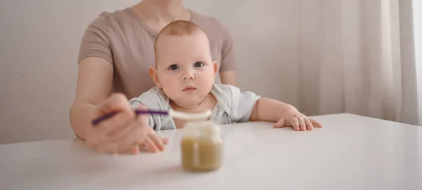 Piccolo neonato divertente bambino che impara a mangiare verdura o purea di frutta da vaso di vetro con cucchiaio. Giovane madre aiutare piccolo figlio mangiare primo cibo. — Foto Stock