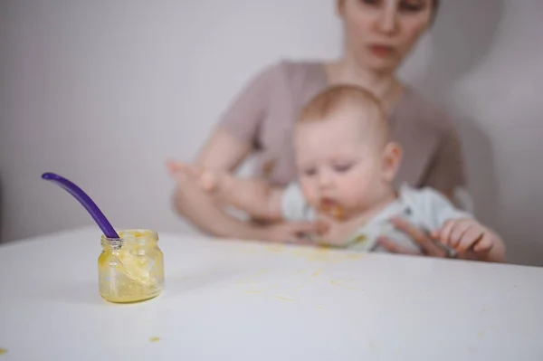 Pequeño bebé divertido recién nacido aprendiendo a comer puré de verduras o frutas de frasco de vidrio con cuchara. Joven madre ayudando a su pequeño hijo a comer primera comida. —  Fotos de Stock