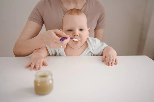 Pequeño bebé divertido recién nacido aprendiendo a comer puré de verduras o frutas de frasco de vidrio con cuchara. Joven madre ayudando a su pequeño hijo a comer primera comida. —  Fotos de Stock