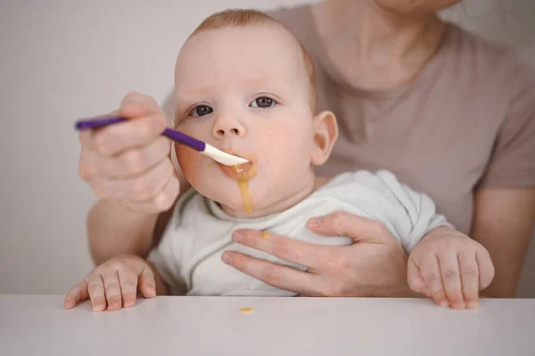 Little newborn funny baby boy learning to eat vegetable or fruit puree from glass jar with spoon. Young mother helping little son eat first food. — Stock Photo, Image