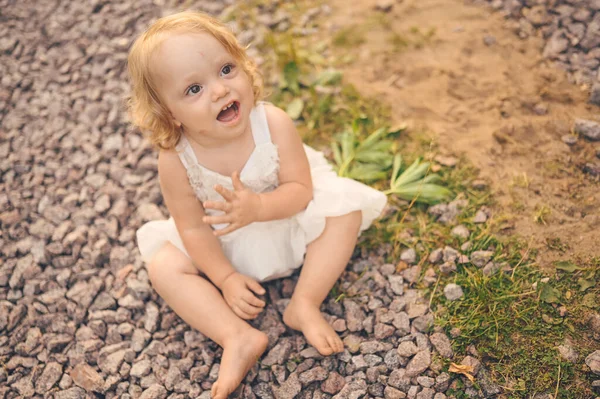 Pequeña niña rubia linda divertida niño pequeño con rizos en vestido blanco y con barro en la cara se sienta en el suelo en la calle fuera en verano. Bebé riendo. Concepto de infancia saludable feliz. —  Fotos de Stock