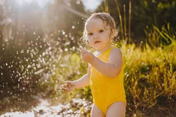 ตลกเล็ก ๆ น้อย ๆ สาวผมบลอนด์น่ารักเด็กวัยหัดเดินในชุดร่างกายเปียกสีเหลืองเล่นริมชายฝั่งริมทะเลสาบที่พระอาทิตย์ตกนอก เด็กที่มีโคลนบนมือและขา กิจกรรมทางน้ําในฤดูร้อน . — ภาพถ่ายสต็อก