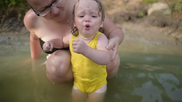 黄色のボディスーツの小さな面白いかわいいブロンドの女の子の子供の幼児は、学習を恐れて泣いて夏の湖で外に泳いでいます。プラスサイズボディ正の女性の母親は天然プールで泳いでいます。子供用タントラ — ストック動画