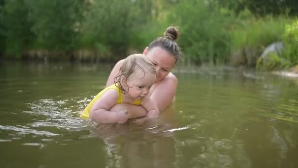 Μικρό αστείο χαριτωμένο ξανθό κορίτσι παιδί νήπιο σε κίτρινο κοστούμι του σώματος μαθαίνει να κολυμπάει έξω στη λίμνη το καλοκαίρι. Συν μέγεθος σώματος θετική χαμογελαστή γυναίκα μητέρα μωρό κολύμπι στη φυσική πισίνα. Πιτσιλιές νερού. — Αρχείο Βίντεο