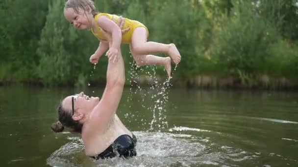 黄色のボディスーツの小さな面白いかわいいブロンドの女の子の子供の幼児笑いは夏の湖で外で泳ぐことを学びます。プラスサイズボディ肯定的な笑顔の女性の母親の赤ちゃんは自然のプールで泳ぐ。水が噴き出す. — ストック動画