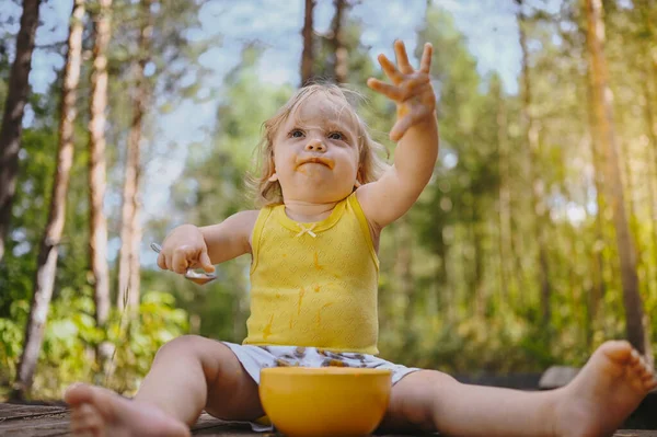 Pouco engraçado bonito menina loira criança criança com roupas sujas e rosto comendo bebê comida fruta ou purê de legumes com colher de prato amarelo fora no verão. Conceito de infância feliz saudável. — Fotografia de Stock