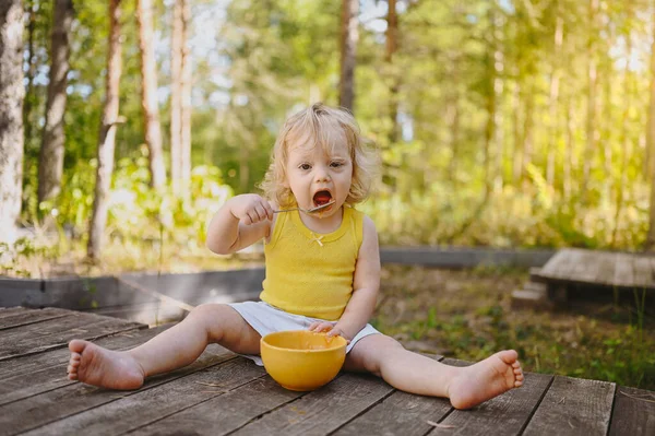 Pequeña niña rubia linda divertida niño pequeño con ropa sucia y la cara comer alimentos para bebés puré de frutas o verduras con cuchara de plato amarillo al aire libre en verano. concepto de infancia feliz saludable. —  Fotos de Stock