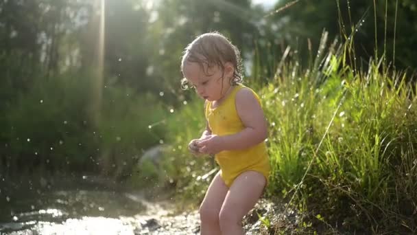 Malá zábavná blondýnka holčička batole ve žlutém mokrém obleku hraje u břehu jezera při západu slunce venku. Dítě s blátem na rukou a nohou. Vodní aktivity v létě. — Stock video
