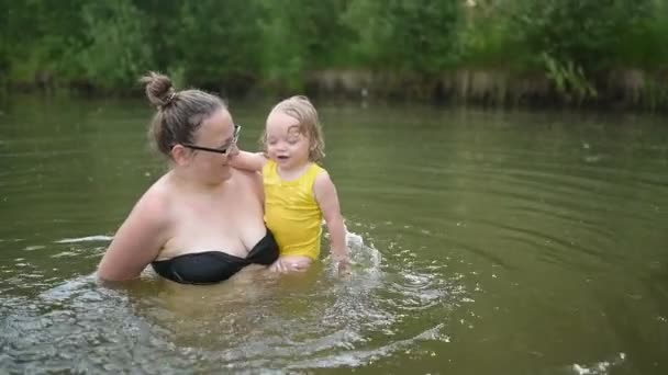 穿着黄色紧身衣笑着的可爱的金发小女孩在夏天的湖中学习游泳。加上身材瘦小、面带微笑的女人妈妈宝宝在自然游泳池里游泳。水花四溅. — 图库视频影像