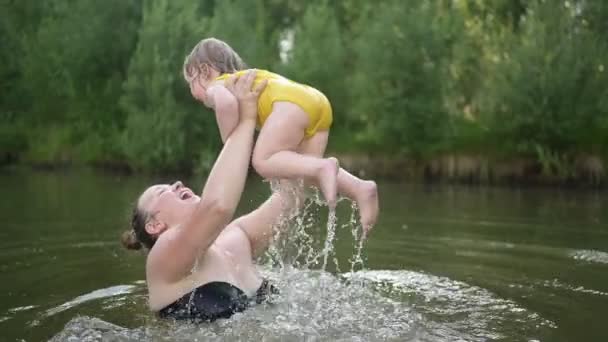 黄色のボディスーツの小さな面白いかわいいブロンドの女の子の子供の幼児笑いは夏の湖で外で泳ぐことを学びます。プラスサイズボディ肯定的な笑顔の女性の母親の赤ちゃんは自然のプールで泳ぐ。水が噴き出す. — ストック動画