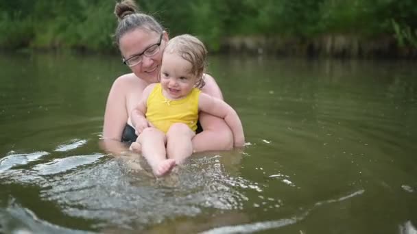 Petite fille blonde mignonne drôle enfant tout-petit en costume jaune riant apprend à nager à l'extérieur au lac d'été. Plus corps taille positive sourire femme mère bébé nager dans la piscine naturelle. Éclaboussures d'eau. — Video