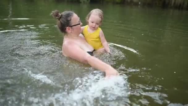 Sarı elbiseli komik sarışın kız çocuğu gülüyor ve yaz gölünde yüzmeyi öğreniyor. Artı vücut ölçüleri pozitif gülümseyen kadın anne bebek doğal havuzda yüzüyor. Su sıçrıyor. — Stok video