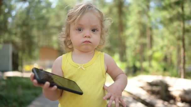 Malá vtipná blondýnka holčička batole drží hrát velký černý smartphone venku v létě na venkově. Zdravý koncept šťastného dětství. Děti a technologie. — Stock video