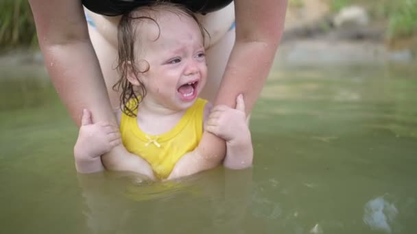黄色のボディスーツの小さな面白いかわいいブロンドの女の子の子供の幼児は、学習を恐れて泣いて夏の湖で外に泳いでいます。プラスサイズボディ正の女性の母親は天然プールで泳いでいます。子供用タントラ — ストック動画