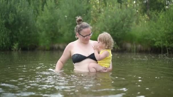 Petite fille blonde mignonne drôle enfant tout-petit en costume jaune riant apprend à nager à l'extérieur au lac d'été. Plus corps taille positive sourire femme mère bébé nager dans la piscine naturelle. Éclaboussures d'eau. — Video