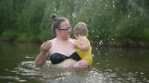 Lilla roliga söta blonda flicka barn småbarn i gul bodysuit skrattar lär simma utanför vid sommarsjön. Plus storlek kropp positiva leende kvinna mor baby simma i naturlig pool. Vattenstänk. — Stockvideo