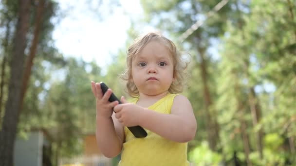 Klein grappig blond meisje kind peuter spelen grote zwarte smartphone buiten in de zomer op het platteland. Gezond concept voor een gelukkige jeugd. Kinderen en technologieën. — Stockvideo