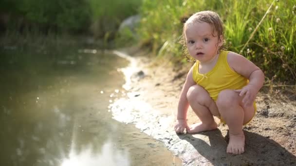 Malá zábavná blondýnka holčička batole ve žlutém mokrém obleku hraje u břehu jezera při západu slunce venku. Dítě s blátem na rukou a nohou. Vodní aktivity v létě. — Stock video