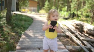 Küçük sevimli sarışın kız çocuğu yazın kırsal kesimde büyük siyah akıllı telefon çalıyor. Sağlıklı mutlu çocukluk kavramı. Çocuklar ve teknolojiler.